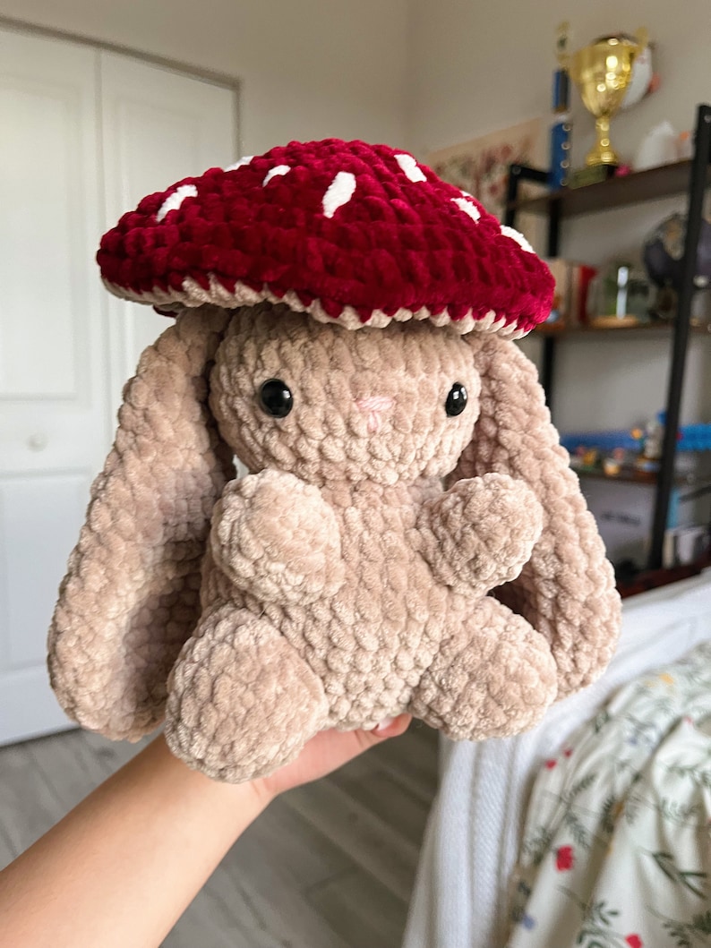 May include: A plush bunny toy with a red mushroom cap with white spots. The bunny is a light brown color with black eyes and a pink nose. The mushroom cap is a deep red color. The toy is soft and cuddly.