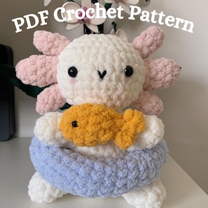 Op de afbeelding: Een handgemaakte gehaakte axolotl-knuffel in wit, roze en blauw, met een gele vis. De tekst "PDF Crochet Pattern" staat erboven. De axolotl heeft zwarte ogen en een lachend gezicht.