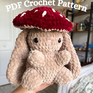 May include: A plush, beige bunny with a red mushroom cap featuring white spots. The bunny has black eyes and a pink nose. The text "PDF Crochet Pattern" is displayed in white across the top of the image. The bunny is soft and cuddly.