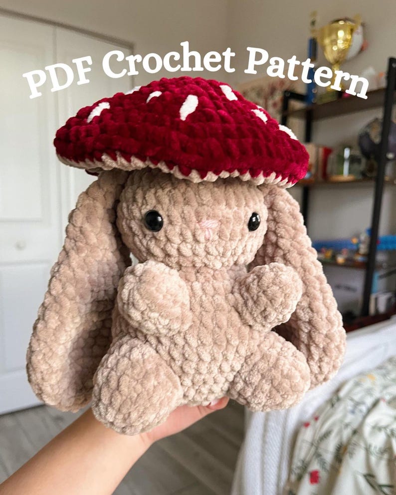 May include: A plush, beige bunny with a red mushroom cap featuring white spots. The bunny has black eyes and a pink nose. The text "PDF Crochet Pattern" is displayed in white across the top of the image. The bunny is soft and cuddly.