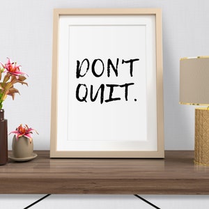 May include: A framed print with the text "DON'T QUIT." in black brush script on a white background. The frame is beige and the print is sitting on a wooden table with a lamp and plants.