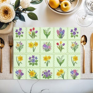 May include: A rectangular placemat with a light green background and a grid pattern. Each square features a different floral illustration, including daffodils, tulips, and violets. Gold-colored cutlery and a white vase with flowers are visible.