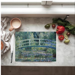 May include: Rectangular cutting board featuring a painting of a bridge over a pond with water lilies, in the style of Claude Monet. The board is surrounded by fresh produce and a mug of coffee.