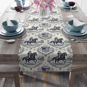 Puede incluir: Camino de mesa color crema con un patrón repetido de figuras ecuestres, banderas americanas y coronas de laurel en azul marino. El camino de mesa está colocado sobre una mesa de madera preparada para una comida, con platos, cuencos y copas de vino.