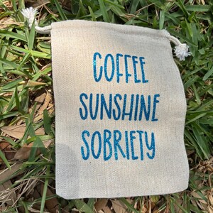 Puede incluir: Una pequeña bolsa de cordón beige con el texto "COFFEE SUNSHINE SOBRIETY" impreso en purpurina azul.