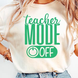 Puede incluir: Camiseta color crema con el texto "teacher MODE OFF" en verde. El diseño incluye un gráfico de botón de encendido/apagado. La camiseta se combina con vaqueros azules.