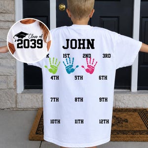以下が含まれることがあります： 黒い文字でJOHNと書かれた白いTシャツ。Kから12THまでの学年がグリッド状に配置されています。K、1ST、2NDの横には緑、青、ピンクの手形があります。インセットには卒業帽とClass of 2039の文字が描かれています。