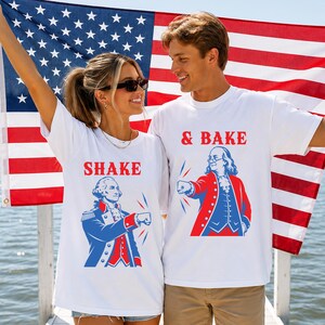 K&ouml;nnte beinhalten: Zwei wei&szlig;e T-Shirts mit rot-blauen Grafikdrucken historischer Pers&ouml;nlichkeiten. Ein Shirt zeigt George Washington mit dem Text SHAKE, das andere Benjamin Franklin mit dem Text & BAKE, beide in einer Faust-an-Faust-Pose.