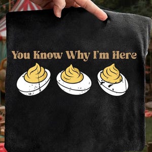 Può includere: Tessuto nero con la scritta "You Know Why I'm Here" sopra tre illustrazioni di uova ripiene. Le uova hanno albumi bianchi e ripieno giallo, con un aspetto invecchiato.