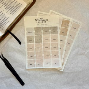 May include: A set of neutral-toned Bible tabs with the text "Wildflower Soiree" and "Mini Bible Tabs." The tabs are arranged in a fan shape, with each tab labeled with a book of the Bible. A pair of black tweezers and an open book are also visible.