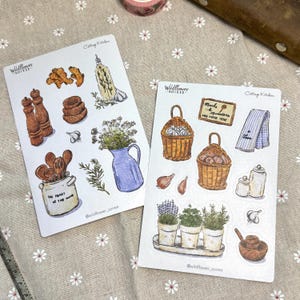 May include: Two sticker sheets with watercolor illustrations of kitchen items. One sheet features a blue pitcher with flowers, wooden spoons, garlic, and spices. The other sheet shows baskets of onions, herbs, and a sign that reads "Meals & Memories Are Made Here."