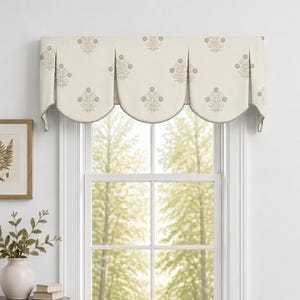 May include: A cream-colored window valance featuring a repeating gray floral pattern with three scalloped sections. The fabric is draped over the top of a window frame, showcasing a structured design with pleats between each rounded panel.