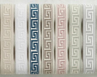 May include: A selection of decorative trim ribbons in various colours, including beige, white, navy blue, grey, and pink. Each ribbon features a Greek key pattern. The ribbons are displayed on a neutral fabric background.