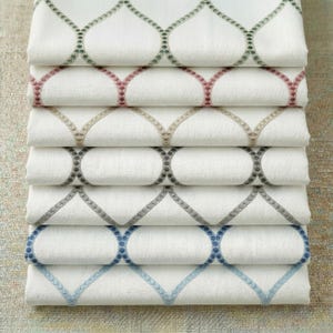 Puede incluir: Una pila de tela blanca con un patrón geométrico bordado en varios colores. Los colores incluyen verde, rosa, beige, gris y azul. El patrón es una serie de óvalos conectados.
