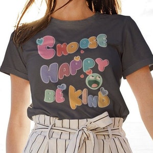 Könnte beinhalten: Ein anthrazitfarbenes T-Shirt mit einer bunten Grafik, auf der "Choose Happy Be Kind" steht. Die Grafik enthält Herzen, Sterne und ein Cartoon-Smiley.