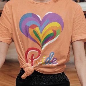 Könnte beinhalten: Ein pfirsichfarbenes T-Shirt mit einem Regenbogenherz-Design und dem Wort "Pride" auf der Vorderseite. Das Shirt ist in der Taille gebunden.
