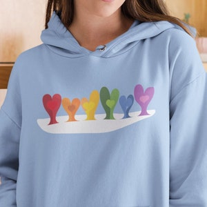 Könnte beinhalten: Ein hellblauer Hoodie mit einem Regenbogen-Herz-Design. Die Herzen sind in einer Reihe auf einer weißen Wolkenform angeordnet.