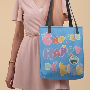 Könnte beinhalten: Eine blaue Tragetasche mit dem Text "Choose Happy Be Kind" in bunten, blasenartigen Buchstaben. Die Tasche hat schwarze Henkel und ein weißes Herzdesign.