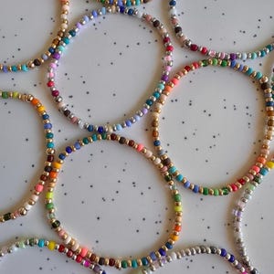 May include: A collection of colorful beaded bracelets. Each bracelet features small, round beads in various colors, including red, orange, yellow, green, blue, and purple. Some bracelets have gold or silver accents. The bracelets are arranged on a white surface.