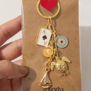 May include: A gold-toned keychain featuring multiple charms attached to a ring, including an ace of spades playing card, a light blue evil eye, a seashell with a pearl, a sea turtle, and a sailboat, all displayed on a brown card with a red heart.