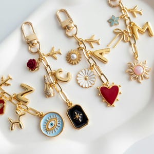 May include: Four gold-toned metal keychains featuring various charms including airplanes, letters, flowers, hearts, suns, a champagne bottle, a balloon dog, an evil eye, and a black rectangular pendant with a star, all attached to lobster clasps.