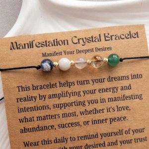 May include: A brown card with the text "Manifestation Crystal Bracelet" and "Manifest Your Deepest Desires". The card shows a black string bracelet with seven beads: a blue and white bead, a white bead, a pink bead, a clear bead with gold flecks, a gold bead, and a green bead.