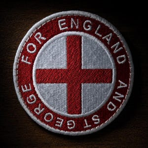 May include: A circular embroidered patch featuring a red cross on a white background. A red border surrounds the centre, containing the white embroidered text "FOR ENGLAND AND ST GEORGE" in a circular arrangement with a white stitched edge.