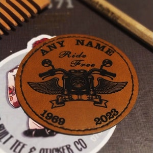 May include: A brown leather patch with black stitching and a motorcycle design. The text "ANY NAME Ride Free" is printed on the patch, along with the years "1969" and "2023".