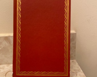 Puede incluir: Un libro de tapa dura rojo con un borde decorativo dorado alrededor de la portada. El libro tiene un marcapáginas de cinta roja.