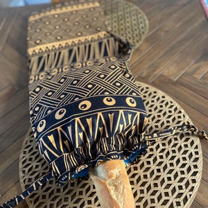 Könnte beinhalten: Eine schwarze Brotbeutel aus Stoff mit goldenen Mustern und einem weißen Baguette darin. Der Beutel liegt auf einer Holzoberfläche mit einem dekorativen, lasergeschnittenen Holzkreis.