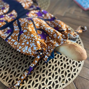 Könnte beinhalten: Eine braune und violette Stoffbrottüte mit einem weißen Muster. Die Tasche ist mit einem Kordelzug verschlossen und ein Laib Brot befindet sich im Inneren.