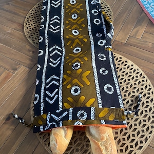Könnte beinhalten: Zwei lange Baguettes sind in einer schwarzen und goldenen Stofftasche mit geometrischem Muster. Die Tasche liegt auf einem Holztisch mit einer geflochtenen Tischunterlage.