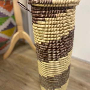 May include: A tall, cylindrical woven basket with a brown and cream pattern. The basket has a lid and a handle.