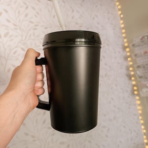 Könnte beinhalten: Ein schwarzer isolierter Becher mit Henkel und Deckel, mit einem weißen, flexiblen Strohhalm. Der Becher wird von einer Hand gehalten, die seine Größe und sein Design zeigt. Der Hintergrund ist eine Wand mit floralem Muster und Lichterketten.