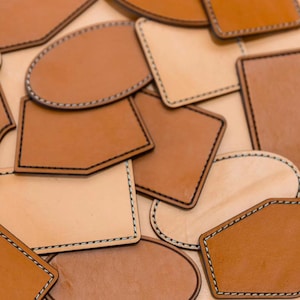 Puede incluir: Un conjunto de parches de cuero de diferentes formas en tonos marrón y beige. Cada parche tiene un borde cosido en negro contrastante. Las formas incluyen rectángulos, óvalos y polígonos irregulares, con un aspecto artesanal.
