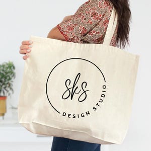 Puede incluir: Bolso tote de color crema con un logotipo circular negro que dice "sks DESIGN STUDIO". El bolso tiene correas largas. El fondo es una pared blanca lisa.