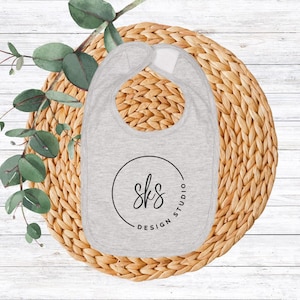 May include: A gray baby bib with a rounded bottom and a neck opening. The bib features a black circular logo with the text "sks DESIGN STUDIO". The bib is placed on a woven, circular placemat with green leaves.