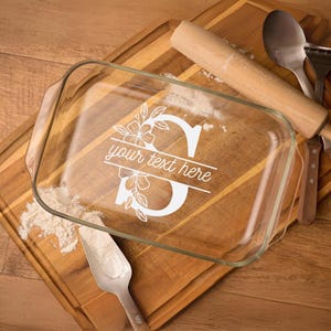 May include: A clear glass baking dish with a white floral design and the letter "S" in the center. The text "your text here" is printed below the letter. The dish is on a wooden cutting board with a rolling pin and a spatula.