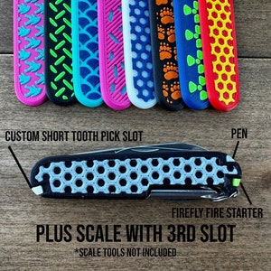 May include: A black and silver multi-tool with a honeycomb pattern on the handle. The tool has a custom short tooth pick slot, a pen, and a firefly fire starter. The text "PLUS SCALE WITH 3RD SLOT" is written below the tool. The text "*SCALE TOOLS NOT INCLUDED" is written below the text.