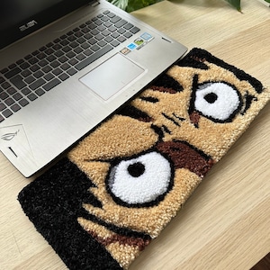 May include: A brown and black rug with a cartoon face on it. The face has large white eyes and a black eyebrow. The rug is on top of a laptop computer.