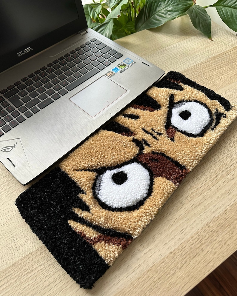 May include: A brown and black rug with a cartoon face on it. The face has large white eyes and a black eyebrow. The rug is on top of a laptop computer.