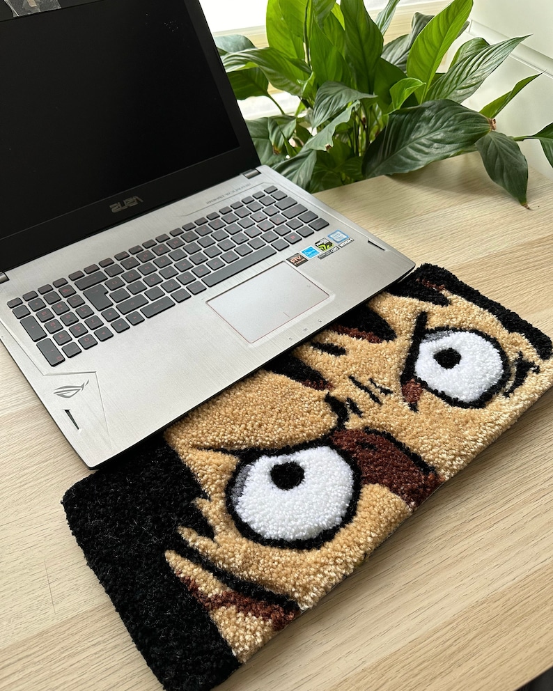 May include: A close-up of a brown and black rug with a cartoon face. The face has large white eyes with black pupils and a brown and black mouth. The rug is partially covered by a silver laptop.