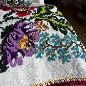 May include: Close-up of a white textile with embroidered floral patterns. The design features large purple flowers with yellow centres, teal leaves, and gold accents. The edge is trimmed with red and yellow crochet.