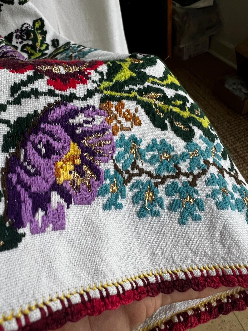 May include: Close-up of a white textile with embroidered floral patterns. The design features large purple flowers with yellow centres, teal leaves, and gold accents. The edge is trimmed with red and yellow crochet.
