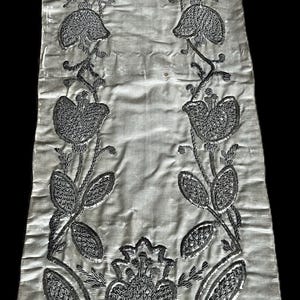 May include: A white fabric runner with silver embroidery of stylized flowers and leaves. The embroidery is detailed and intricate, creating a delicate and elegant design.
