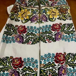 May include: White embroidered garment with colourful floral patterns. The design includes red roses, yellow and purple flowers, and green leaves. The embroidery is detailed and covers the front of the garment.
