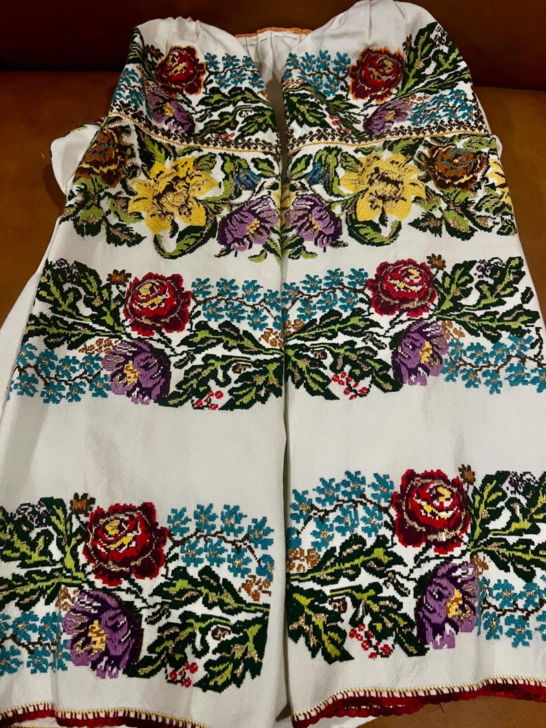 May include: White embroidered garment with colourful floral patterns. The design includes red roses, yellow and purple flowers, and green leaves. The embroidery is detailed and covers the front of the garment.