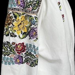 May include: White blouse with intricate floral embroidery in shades of red, yellow, purple, and green. The sleeves are heavily decorated with the embroidered design. The blouse has a round neck and long sleeves.