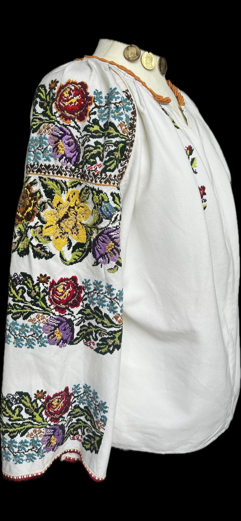 May include: White blouse with intricate floral embroidery in shades of red, yellow, purple, and green. The sleeves are heavily decorated with the embroidered design. The blouse has a round neck and long sleeves.