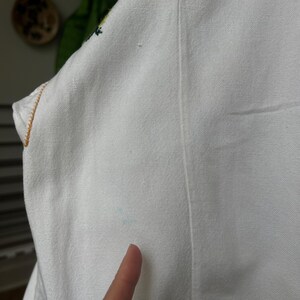 May include: Close-up of a white fabric garment with a visible seam and a small area of blue discoloration. The edge of the garment is trimmed with a gold-coloured thread. The fabric appears to be a woven material.